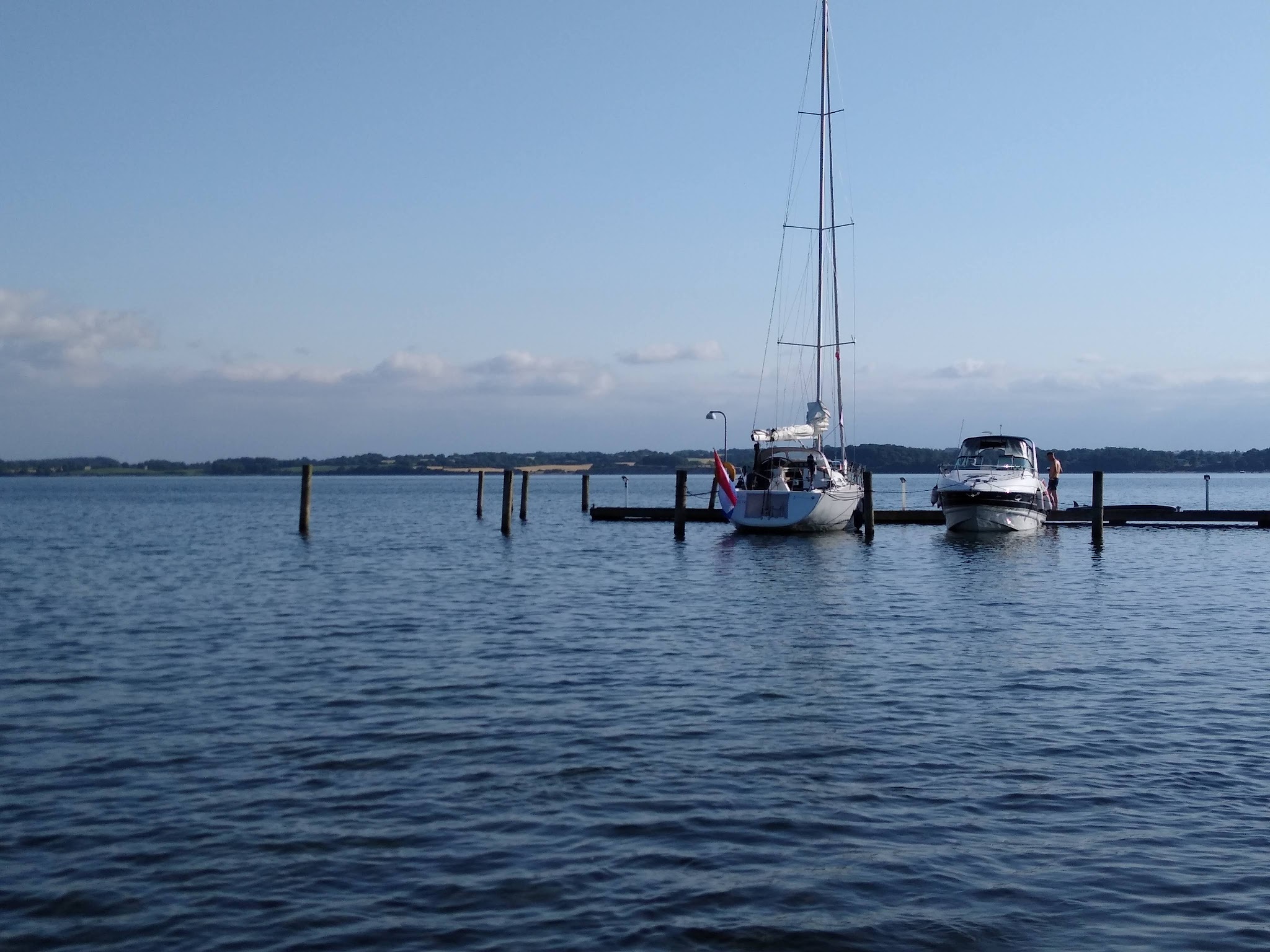 Zwei Boote liegen an kleinem Steg auf ruhigem See, blauer Himmel und ferne Uferlinie im Hintergrund.