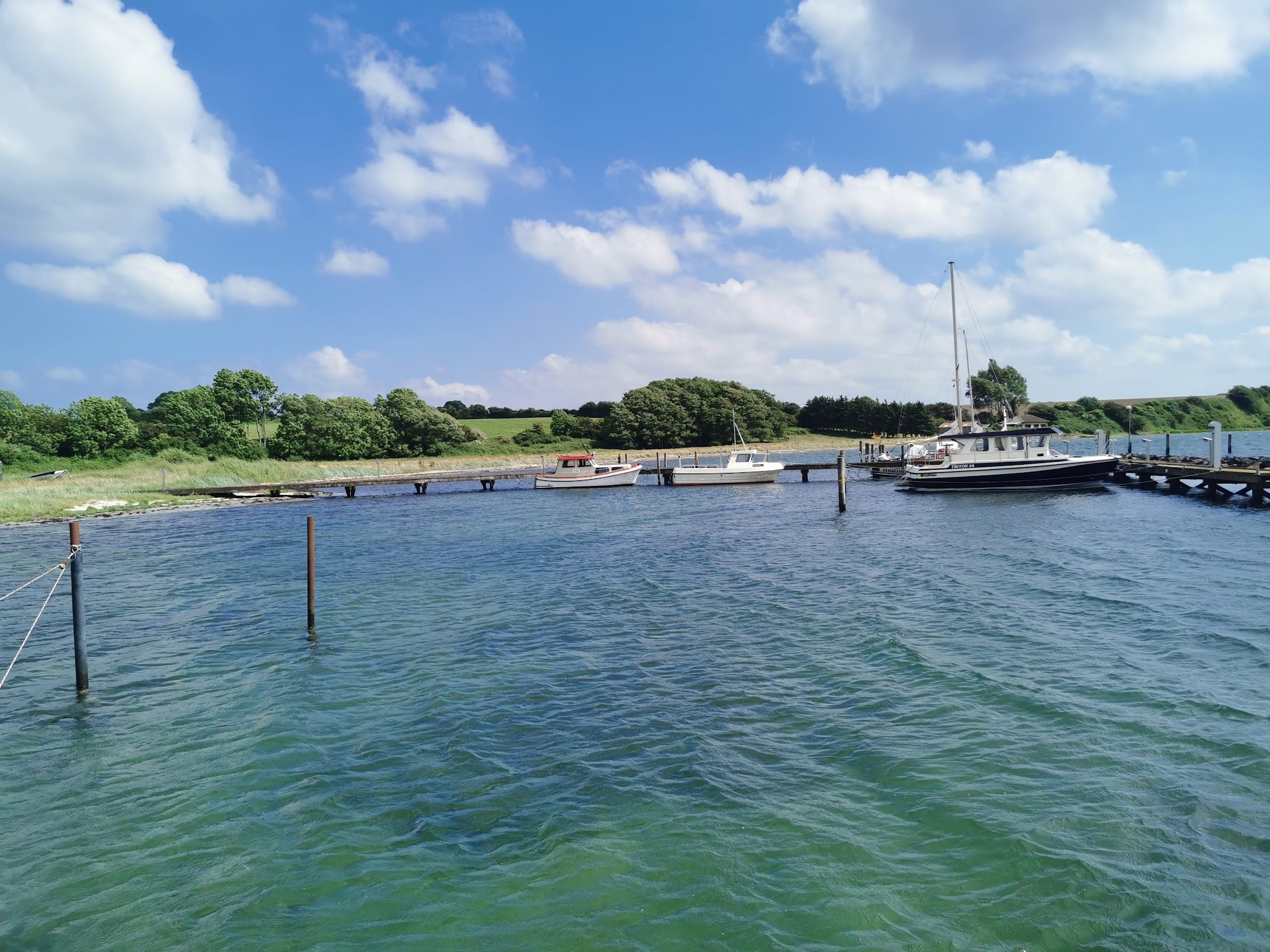 Kleiner Anleger mit Segel- und Motorbooten, grünes Ufer, blaues Wasser unter leicht bewölktem Sommerhimmel