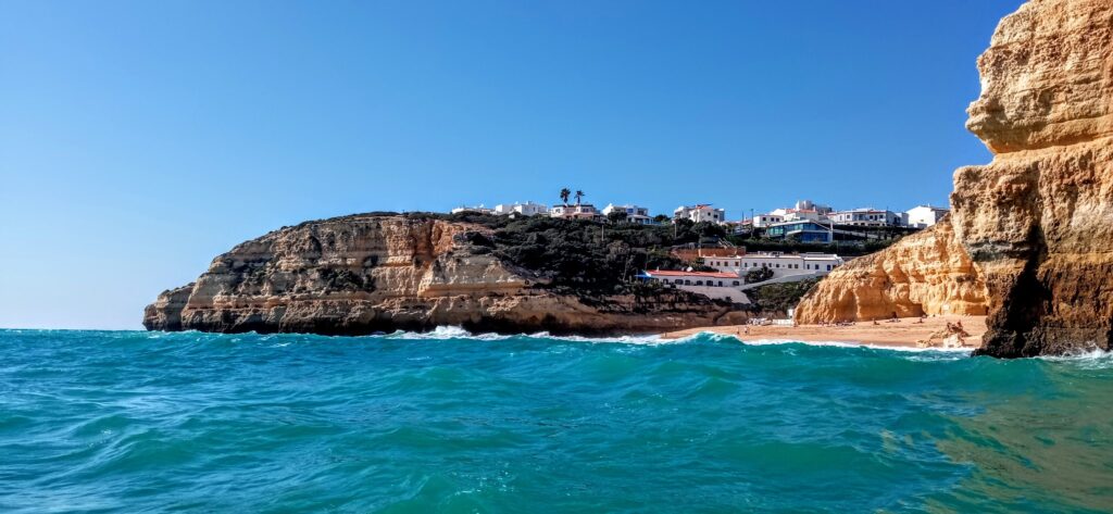 Türkisblaues Meer vor gestreiften Kalkklippen, kleiner Sandstrand mit wenigen Leuten, weiße Häuser auf dem Hügel