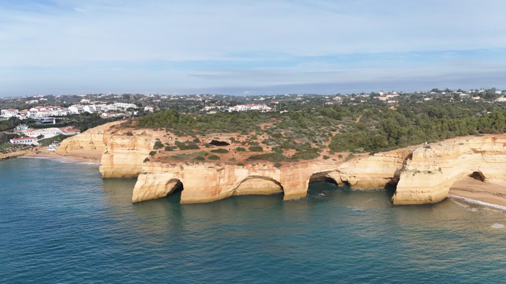 Gelbe Felsklippen mit drei Meeresbögen ragen ins türkisfarbene Wasser; dahinter grünes Hügelland und weiße Küstenhäuser.