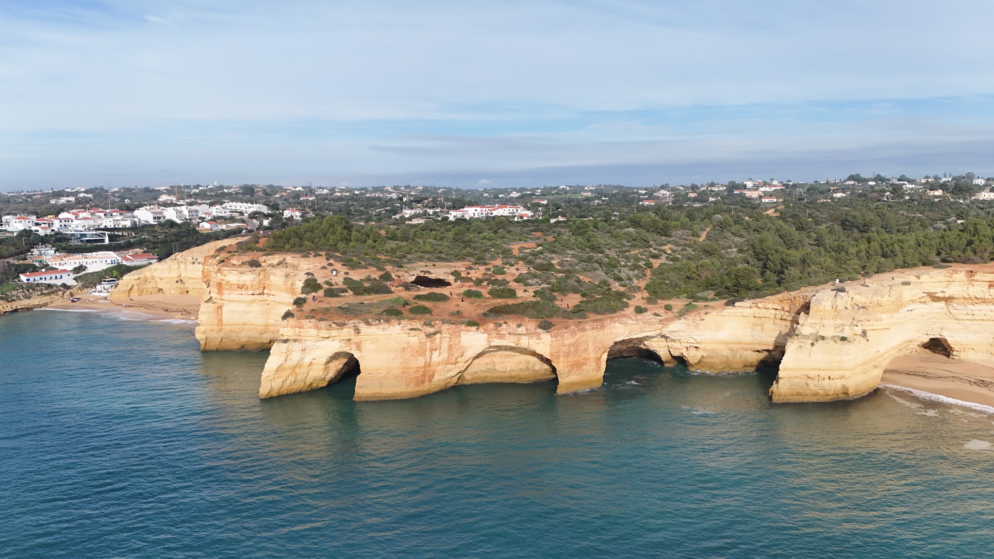Gelbe Felsklippen mit drei Meeresbögen ragen ins türkisfarbene Wasser; dahinter grünes Hügelland und weiße Küstenhäuser.