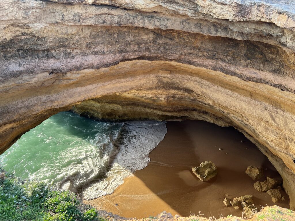 Blick von oben in eine Felsgrotte: türkisgrünes Meer brandet auf sandigen Strand, Sonnenlicht und Felsbrocken.