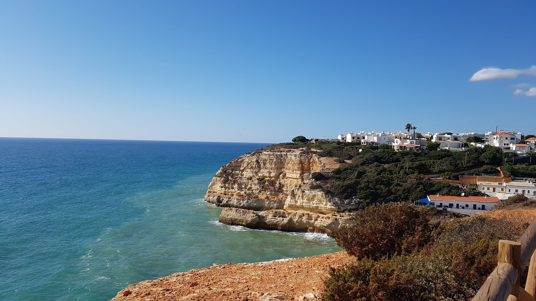 Steilküste mit türkisblauem Meer, gelblichen Felswänden und weißen Häusern auf dem begrünten Plateau unter klarem Himmel.