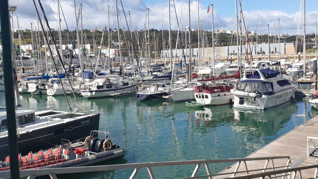 Dicht aneinandergereihte Segelboote und Yachten im sonnigen Hafen; Masten spiegeln sich im türkisgrünen Wasser.