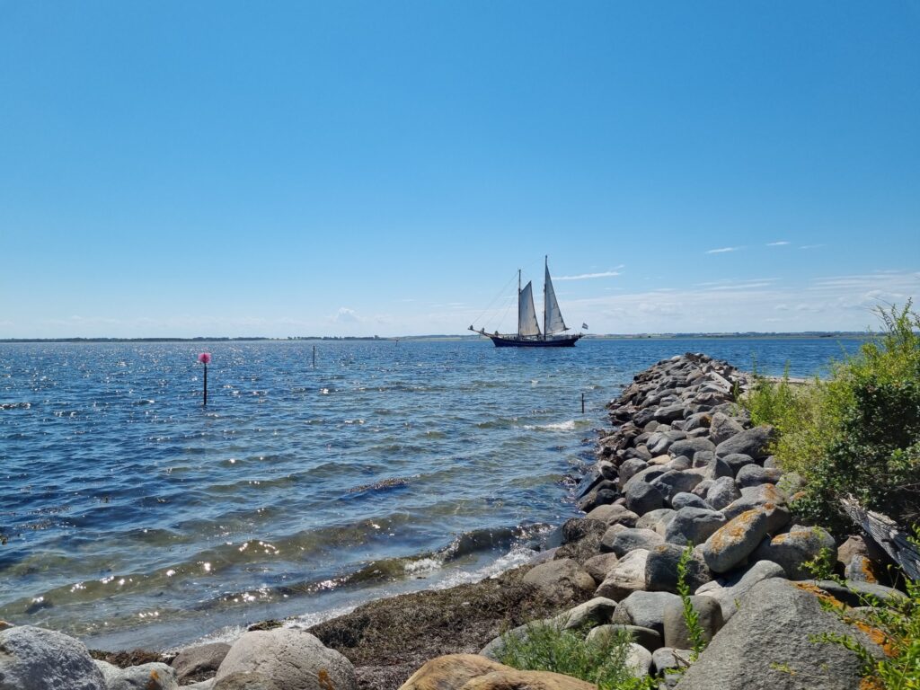 Segelschiff auf glitzerndem Meer, vorbei an steinigem Wellenbrecher unter wolkenlosem, blauem Himmel