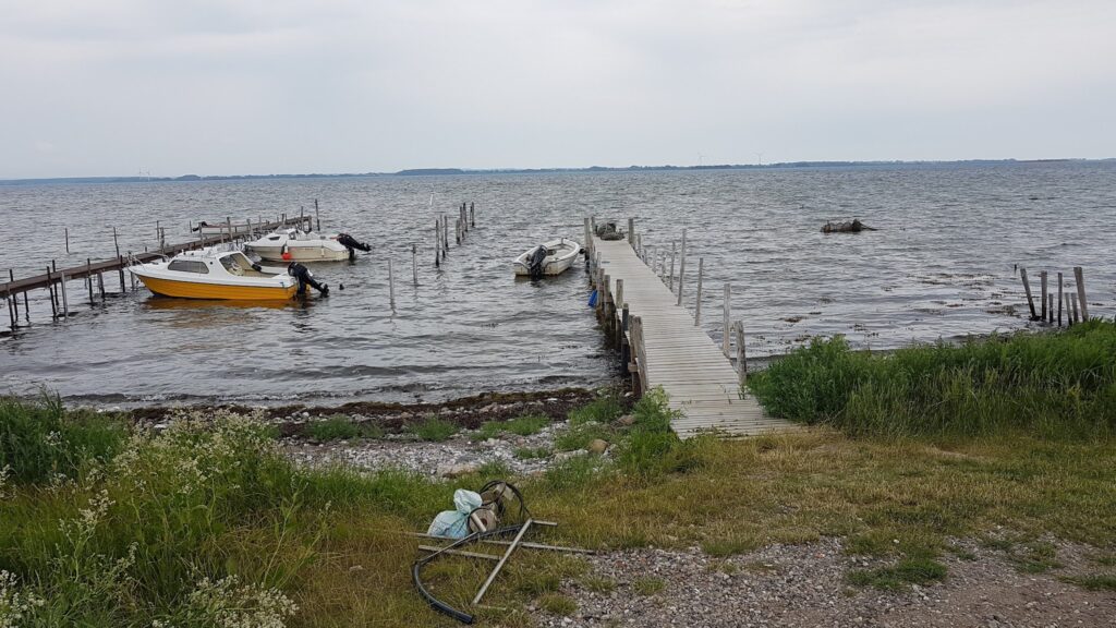 Holzsteg ragt in graue, windgekräuselte See; gelbe und weiße Motorboote vertäut, grasiger Kiesstrand im Vordergrund
