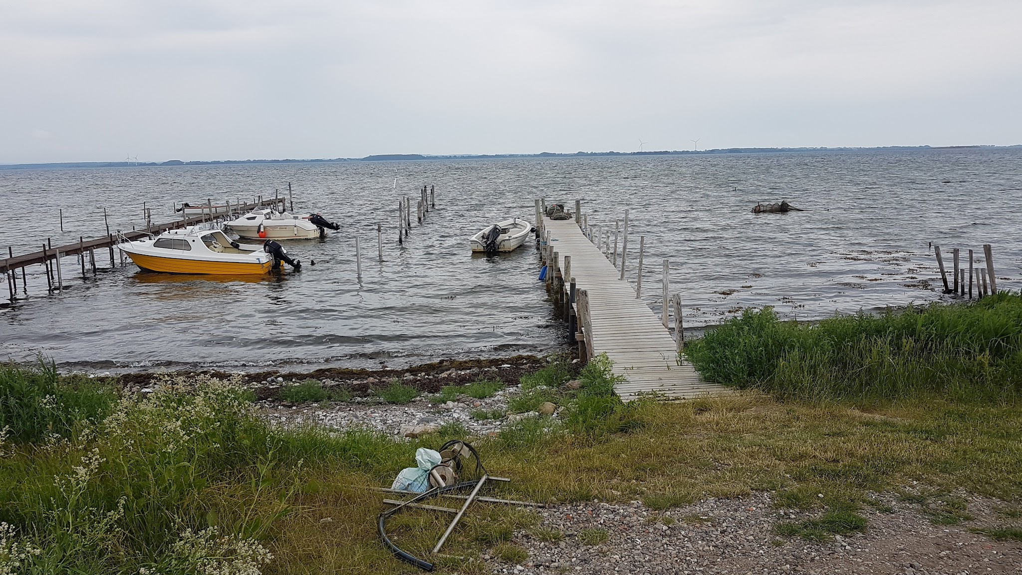 Holzsteg ragt in graue, windgekräuselte See; gelbe und weiße Motorboote vertäut, grasiger Kiesstrand im Vordergrund