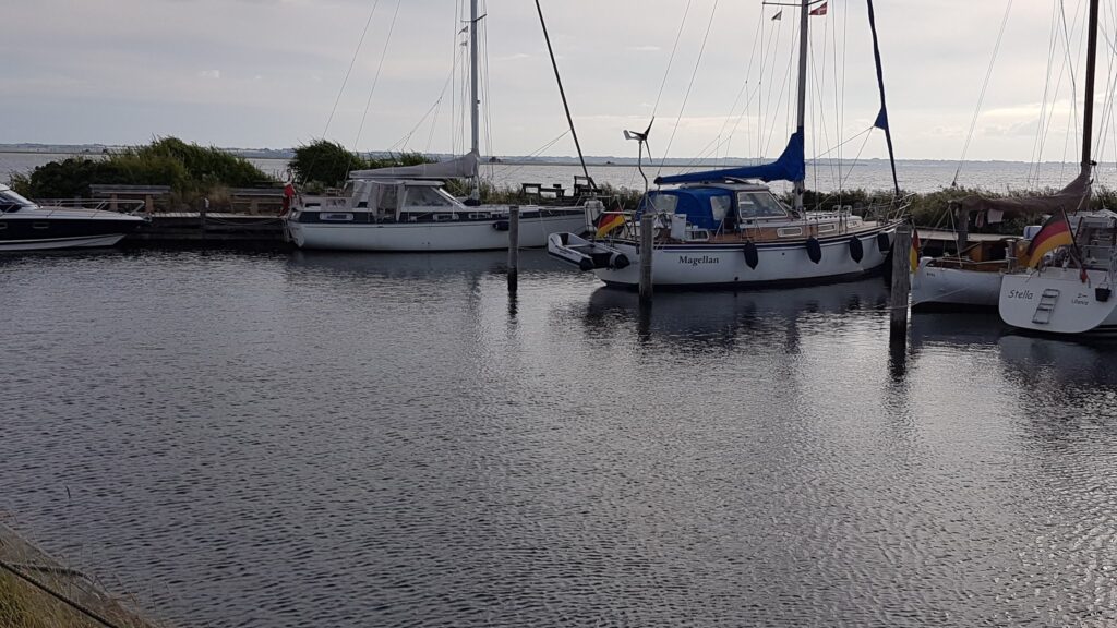 Mehrere Segelboote mit deutscher Flagge liegen still im kleinen Hafen; ruhiges, leicht bewölktes Küstenpanorama.