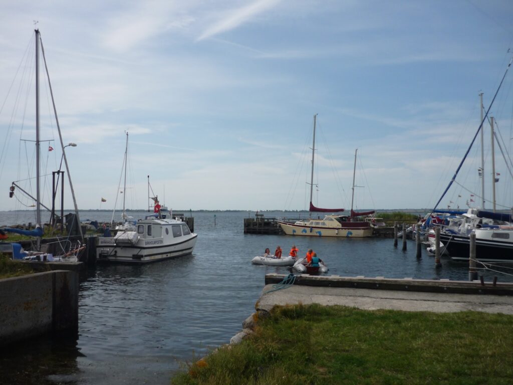 Kleiner Hafen, Segelboote am Steg, Kinder in orangen Rettungswesten paddeln im Schlauchboot auf ruhigem Wasser.