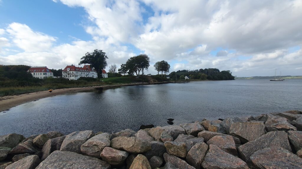 Steinmole am ruhigen Fjord; weiße Häuser mit roten Dächern, Bäume und segelndes Boot unter leicht bewölktem Himmel