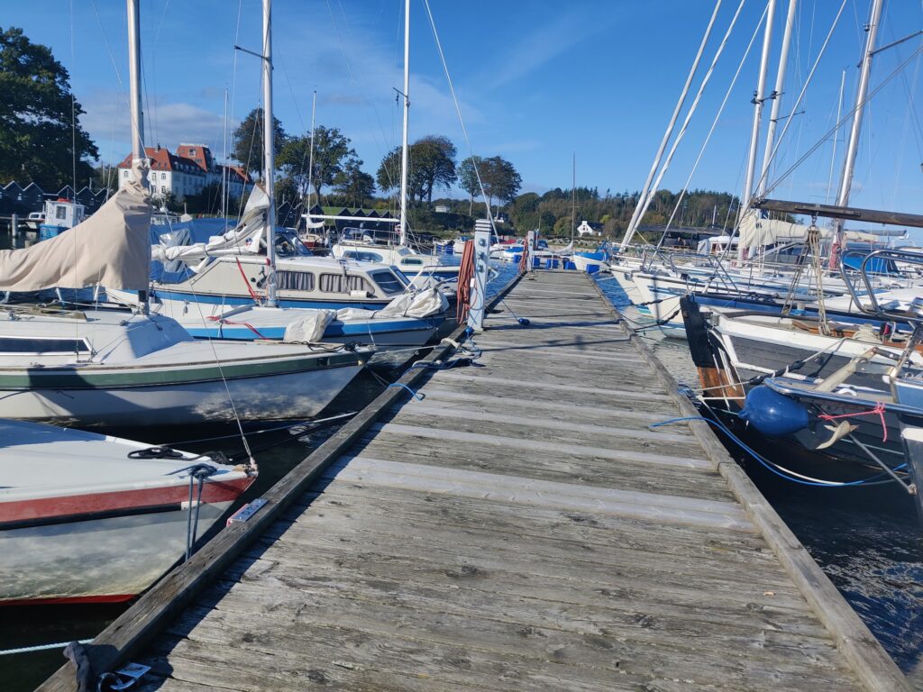 Holzsteg mit vertäuten Segelbooten, ruhiger Hafen unter blauem Himmel, rotdachige Villa im Hintergrund