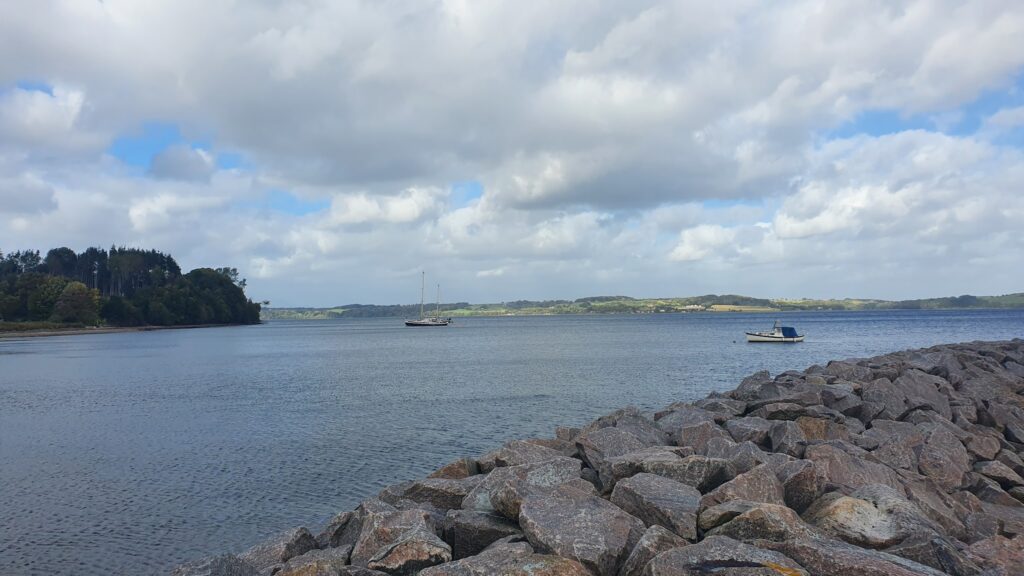 Ruhige Bucht mit Felsufer, Segelboot mitten im Wasser, kleines Motorboot rechts, bewaldete Landzunge im Hintergrund