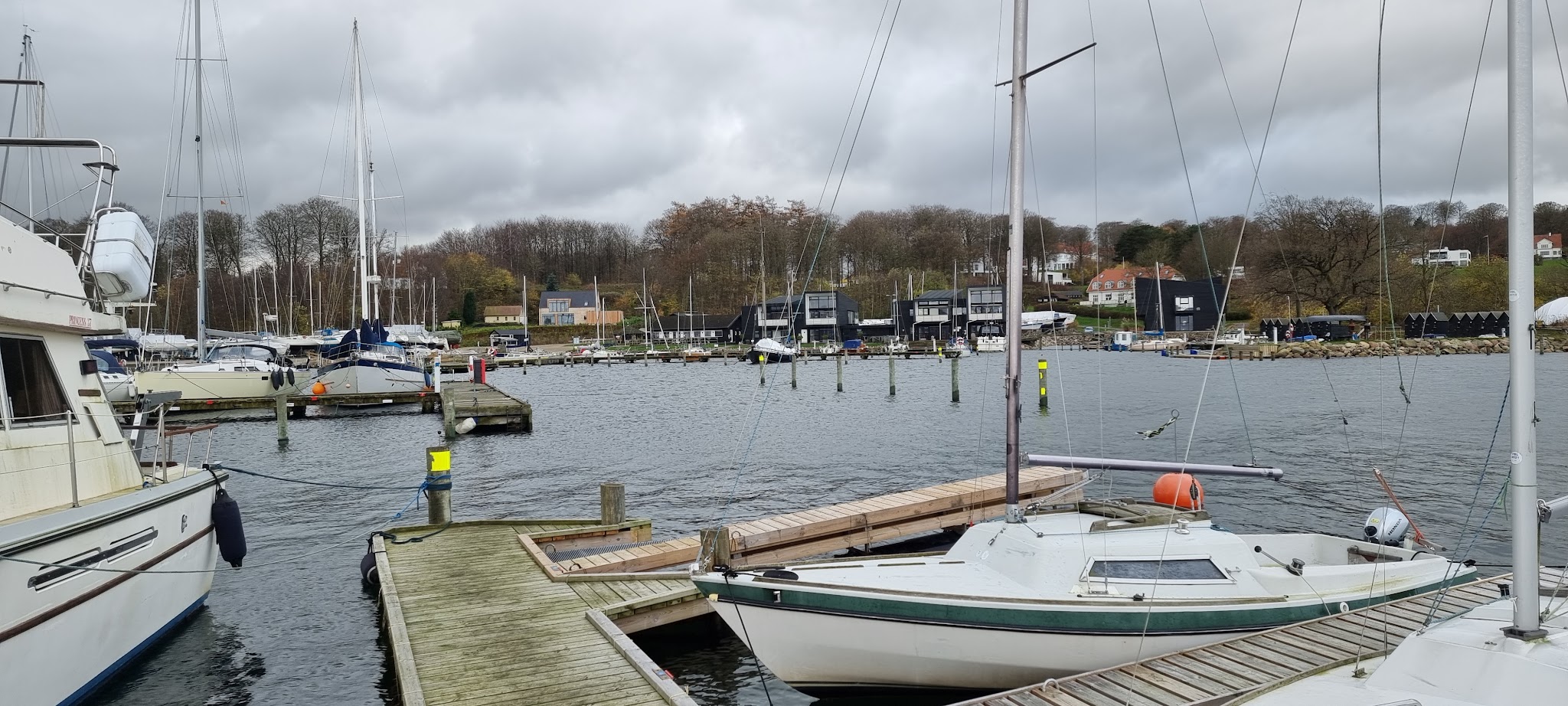 Bewölkter Jachthafen: Segel- und Motorboote an Holzstegen, dahinter Uferhäuser und herbstliche Bäume.