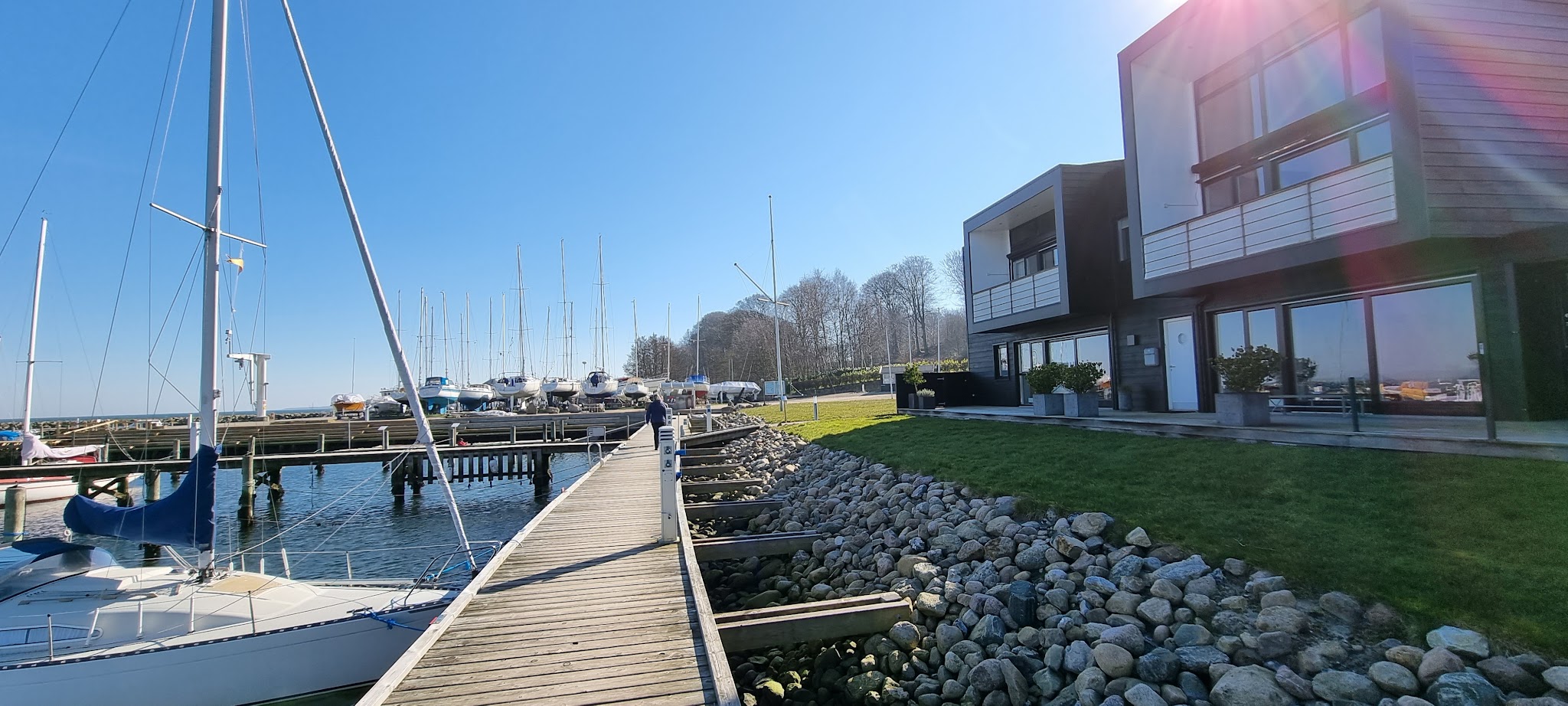 Holzsteg mit Segelbooten im Yachthafen; Person geht Richtung an Land gelagerter Boote, moderne Uferhäuser rechts.