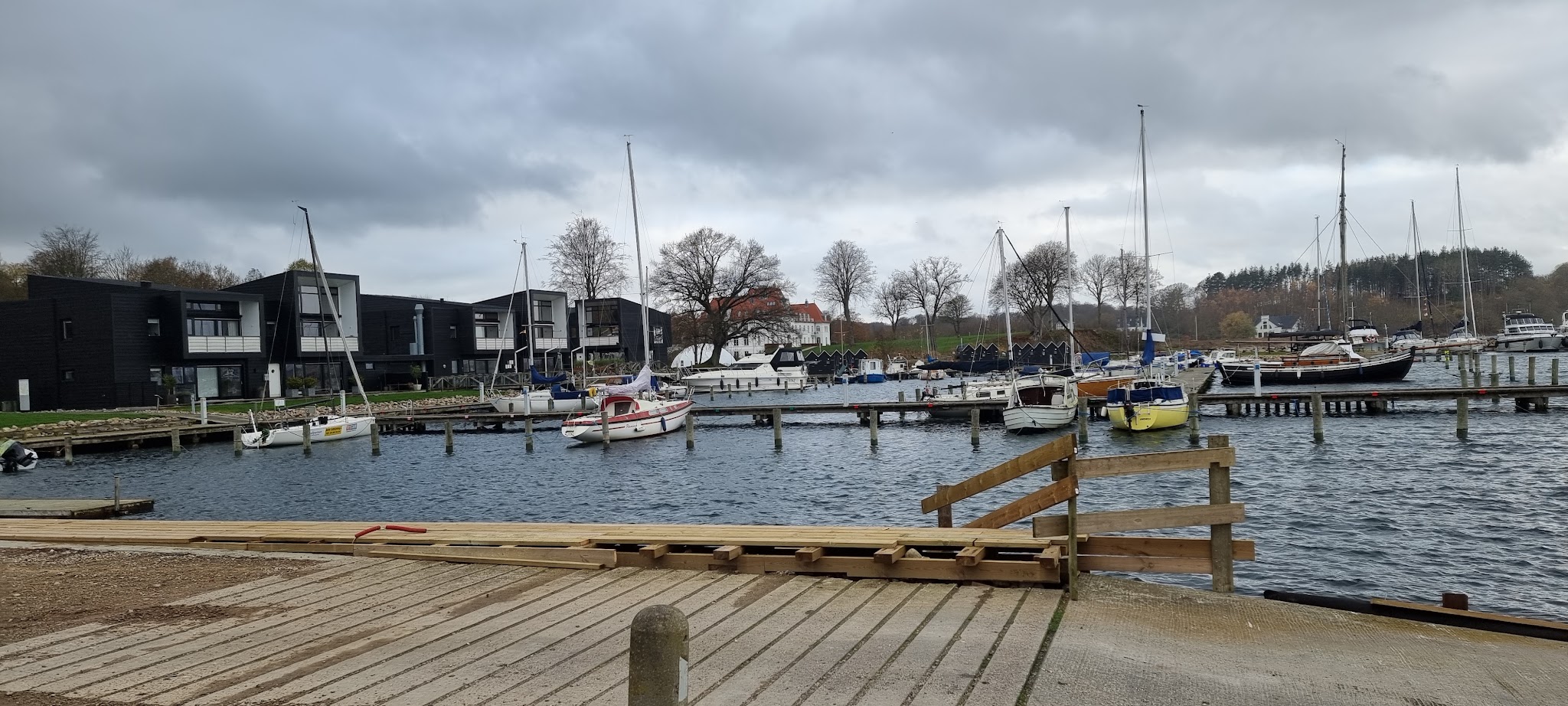 Kleiner Yachthafen mit mehreren Segelbooten vor modernen schwarzen Häusern unter bewölktem Himmel.