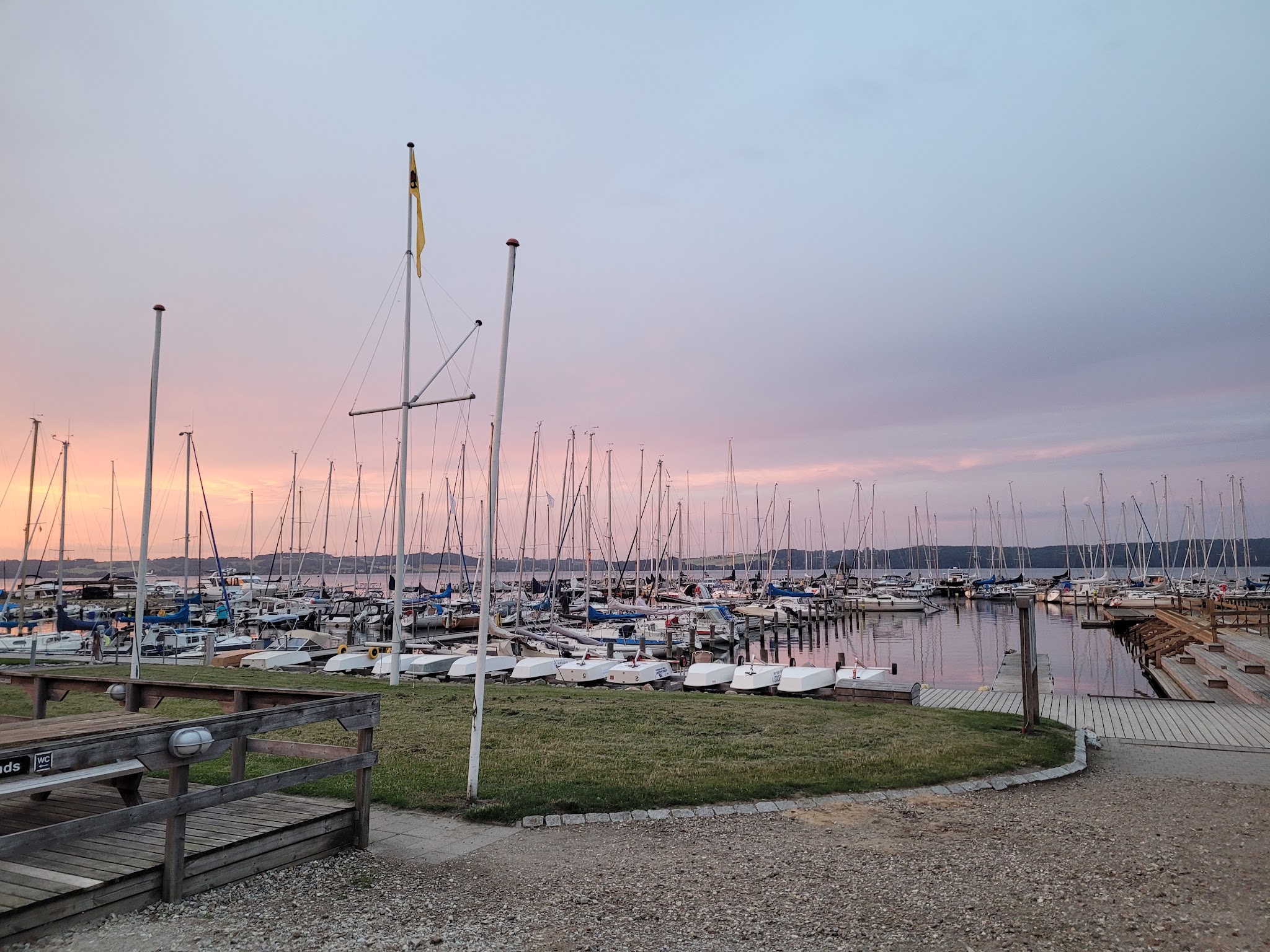 Sonnenuntergang über einem Yachthafen; zahlreiche Segelboote mit aufragenden Masten liegen ruhig an den Stegen.