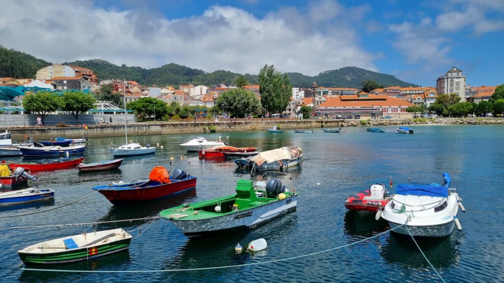 Kleine bunte Boote liegen im ruhigen Hafenwasser; dahinter Küstenstadt mit grünen Hügeln unter teils bewölktem Himmel