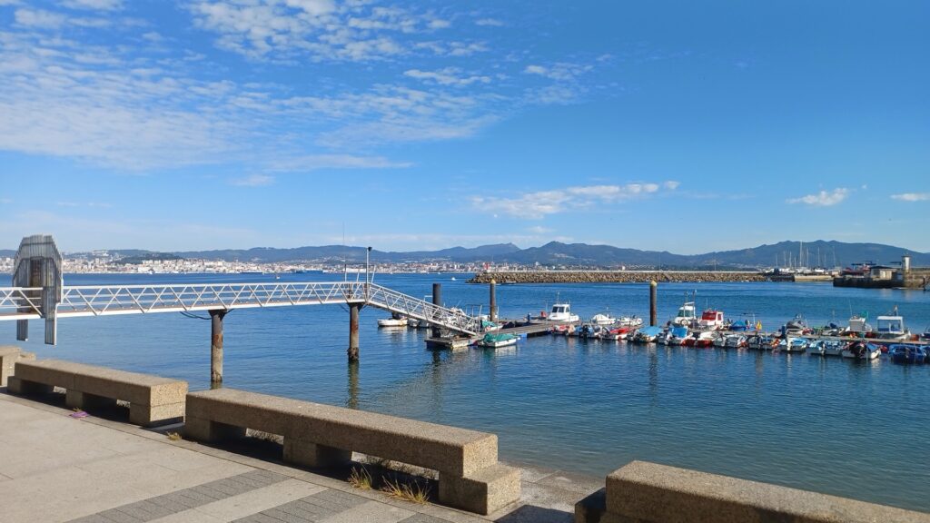 Kleiner Bootssteg mit vielen bunten Fischerbooten vor ruhiger Bucht; Promenade, Stadt und Berge im Hintergrund