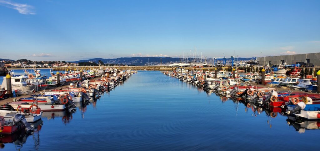 Reihen bunter Fischerboote liegen beidseits eines ruhigen Hafenbeckens, Himmel und Berge spiegeln sich im Wasser.