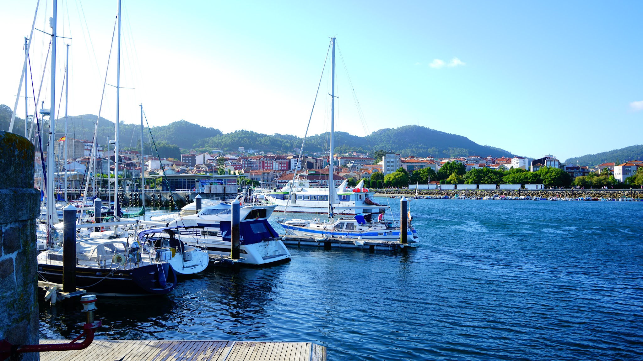 Jachthafen mit Segelbooten auf blauem Wasser; dahinter Küstenstadt vor grünen Hügeln unter klarem Himmel