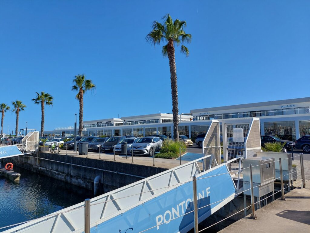 Rampe mit Schild „PONTON F“ am Hafen; dahinter geparkte Autos, Palmen und modernes, flaches Gebäude im Sonnenschein.