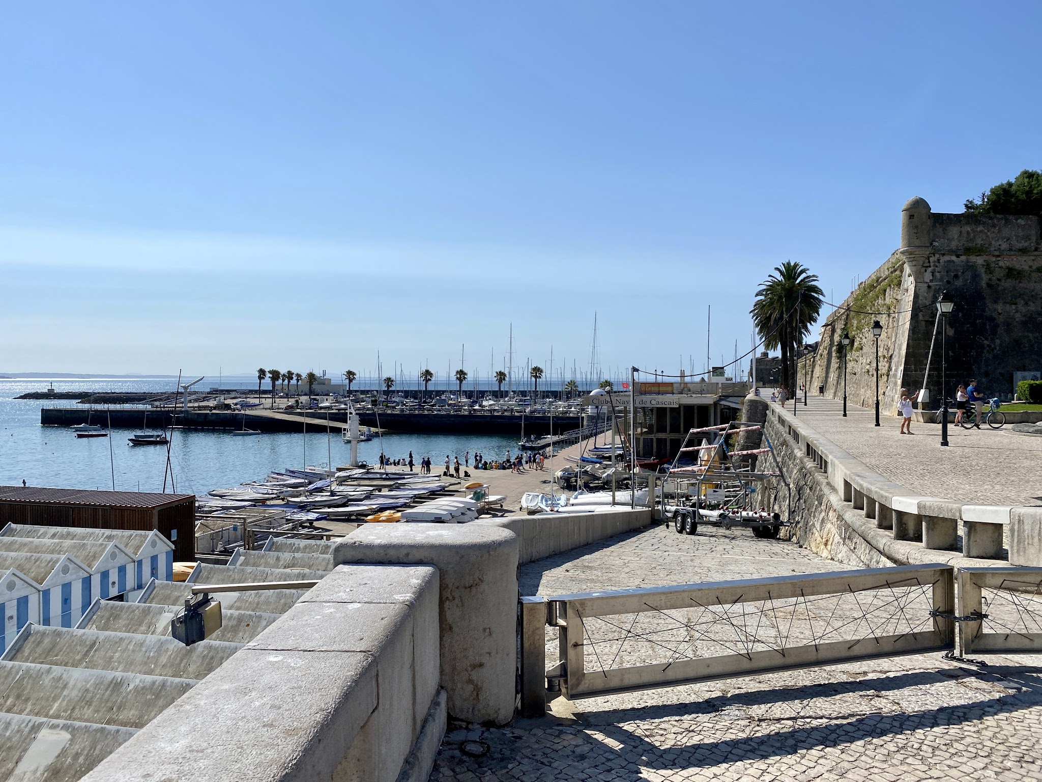 Sonniger Hafen mit Segelbooten, Palmen und Festungsmauer; Spaziergänger auf der Steinpromenade.