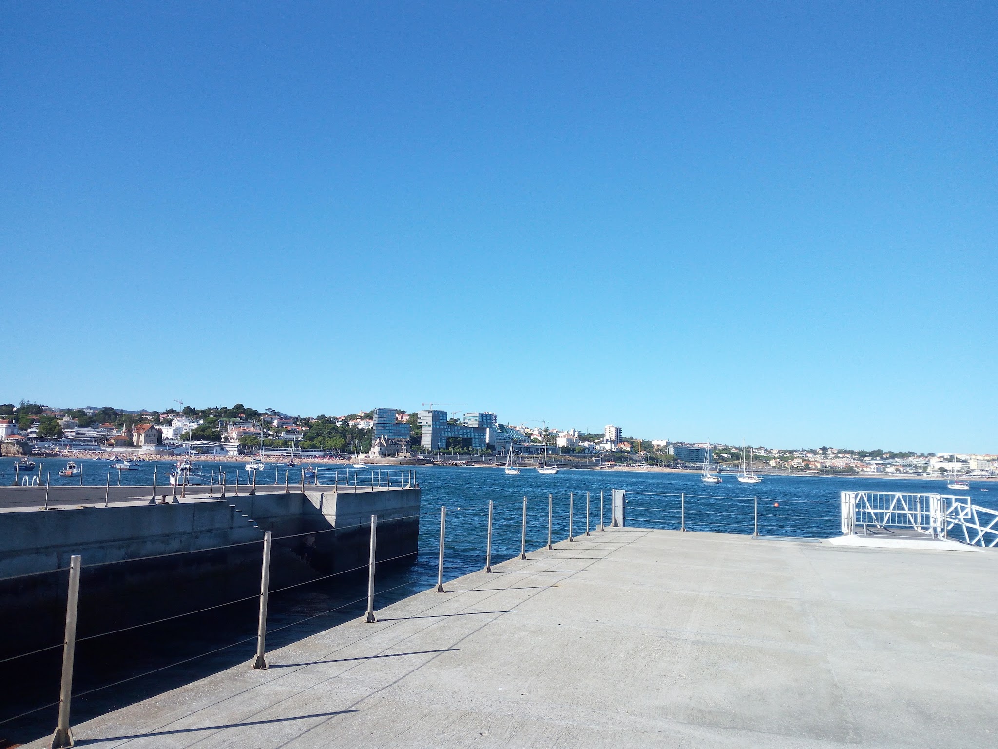 Breiter Betonsteg mit Geländer, vorne Meerbucht mit mehreren Segelbooten, dahinter Küstenstadt unter wolkenlosem Himmel