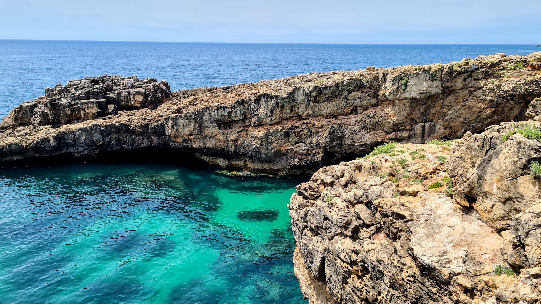 Türkis schimmernde Meeresbucht unter zerklüfteten, hellbraunen Felsklippen, dahinter offenes, ruhiges Meer
