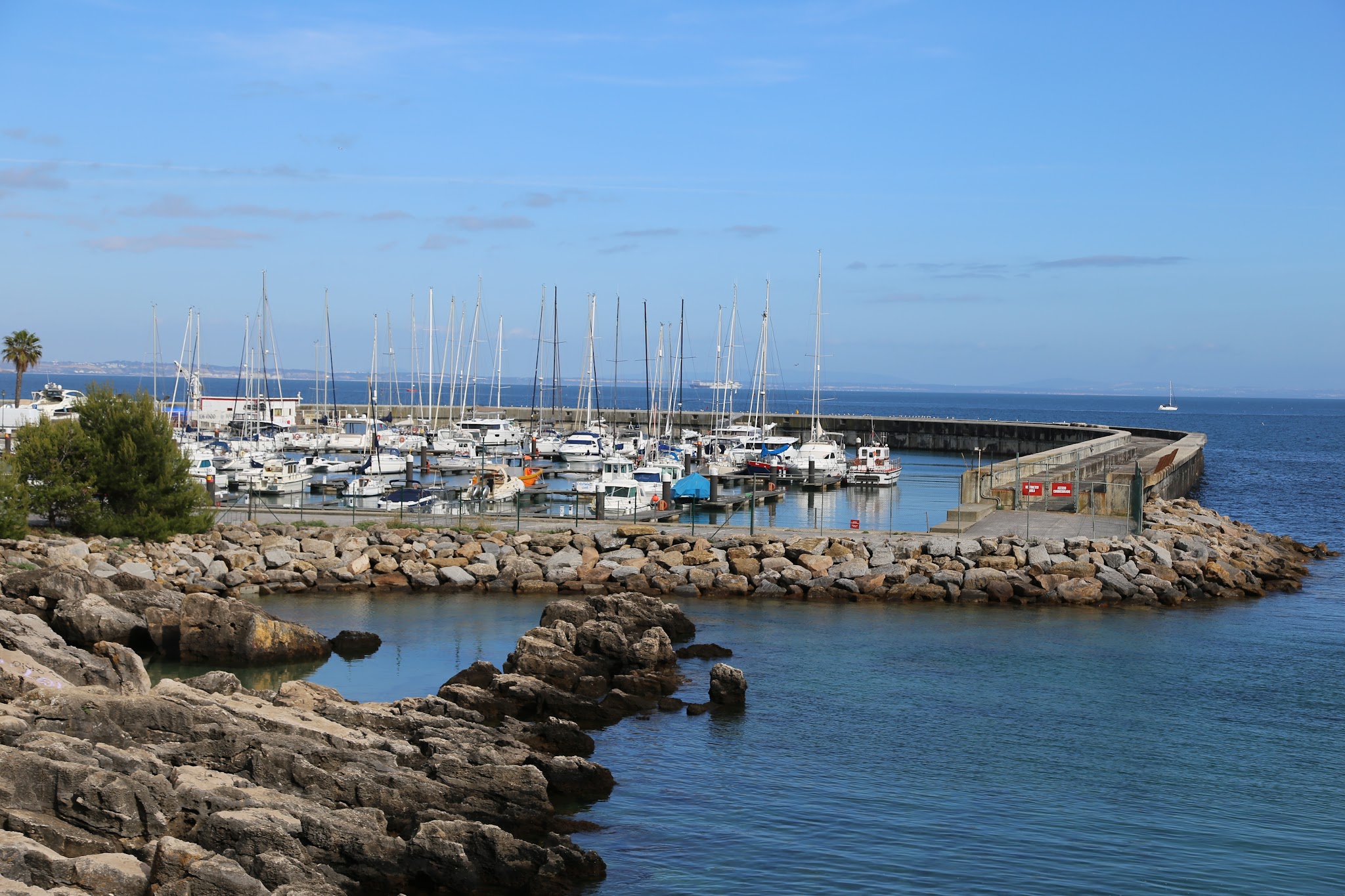 Marina mit Segelbooten, umgeben von Felsmole und felsiger Küste; ruhiges blaues Wasser unter wolkenlosem Himmel.