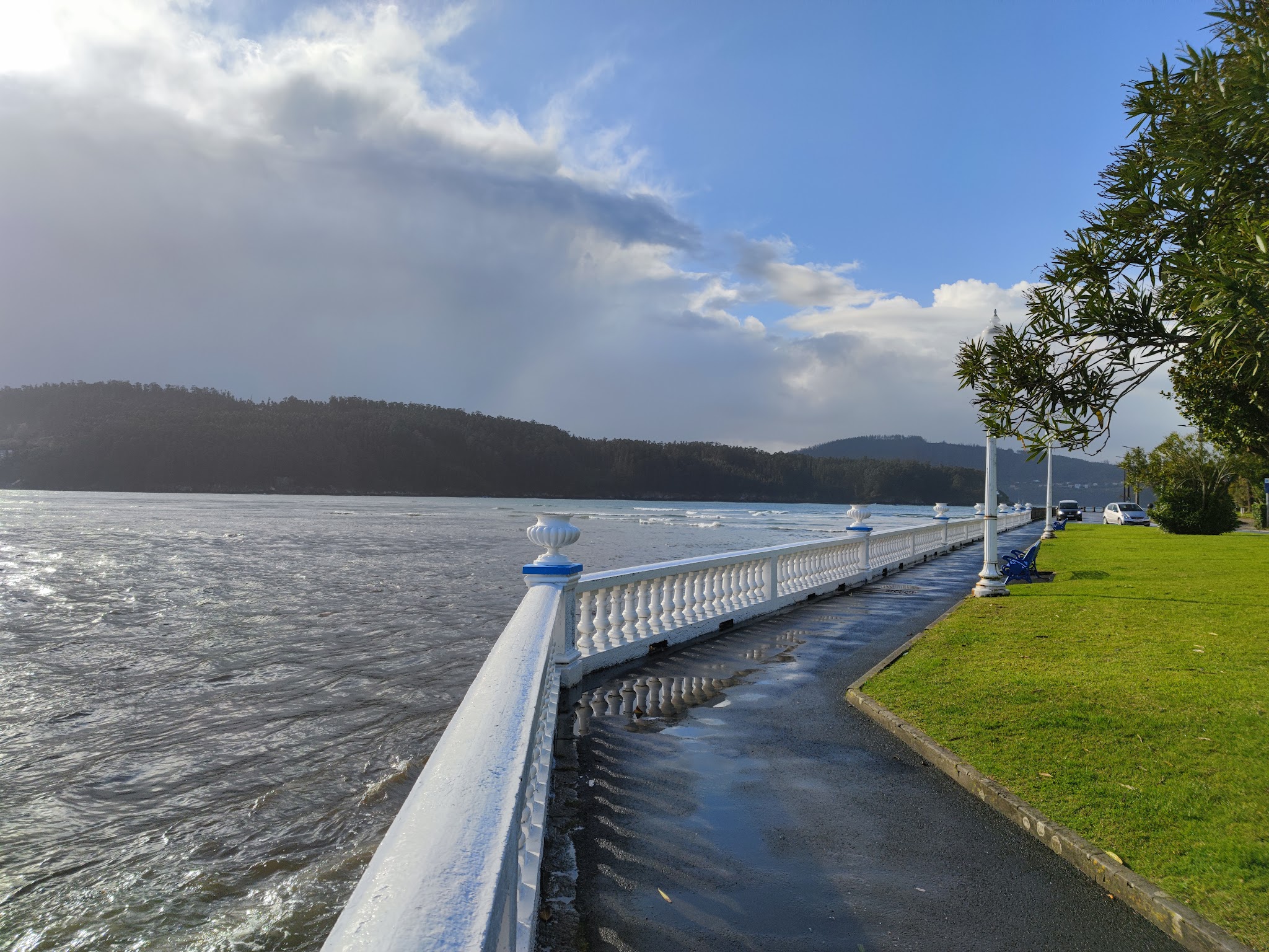 Nasse Küstenpromenade mit weißer Balustrade, grünem Rasen, ruhigem Meer und bewaldeten Hügeln unter wechselhaftem Himmel.