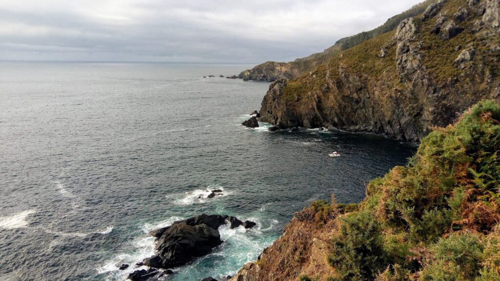 Steile Felsküste mit grünem Bewuchs; unten brandet dunkelblaues Meer, ein kleines weißes Boot ankert in der Bucht.
