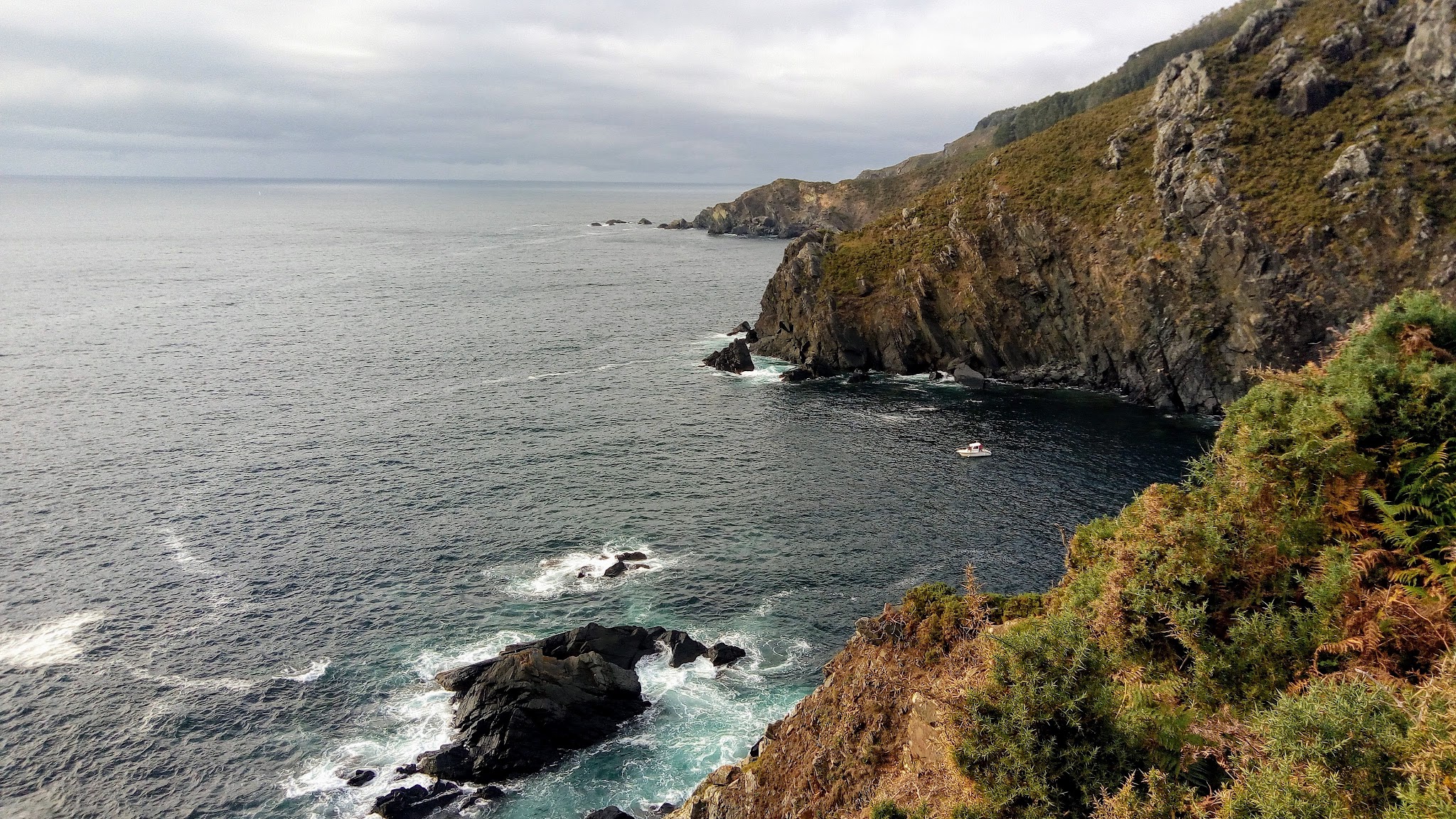 Steile Felsküste mit grünem Bewuchs; unten brandet dunkelblaues Meer, ein kleines weißes Boot ankert in der Bucht.