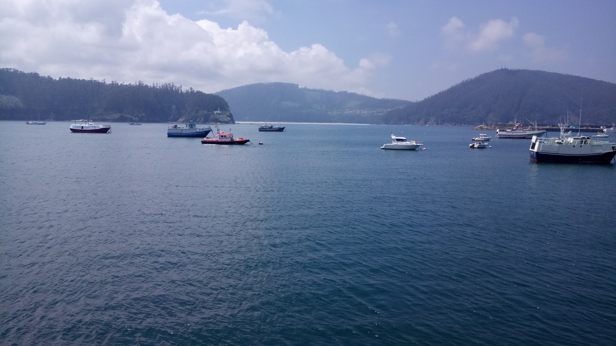 Mehrere Boote liegen in ruhiger, blauer Bucht; bewaldete Hügel und leichter Wolkenhimmel im Hintergrund.