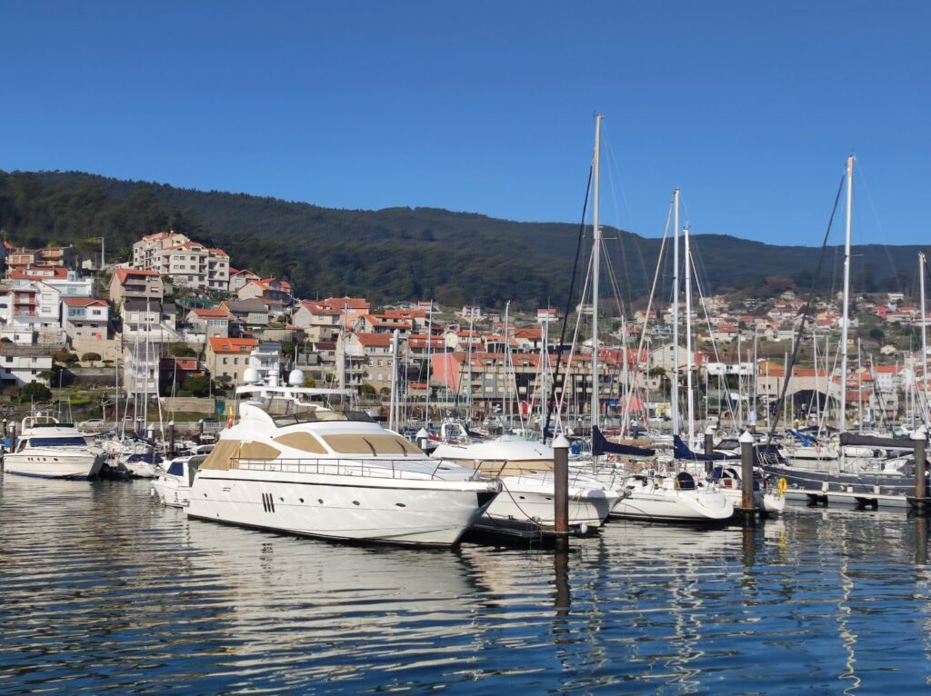 Yachten und Segelboote im Hafen; dahinter Hang mit Häusern mit roten Dächern und bewaldetem Hügel unter klarem Himmel