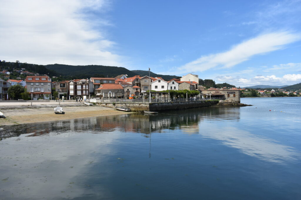 Kleine Küstenstadt mit roten Ziegeldächern an ruhiger Bucht; Boote am flachen Ufer, bewaldete Hügel im Hintergrund