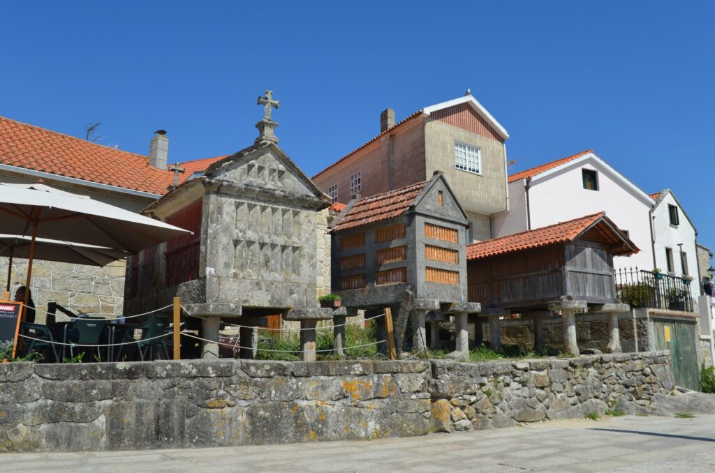 Steinerne und hölzerne galicische Hórreos auf Pfeilern vor Häusern mit roten Ziegeldächern unter blauem Himmel.