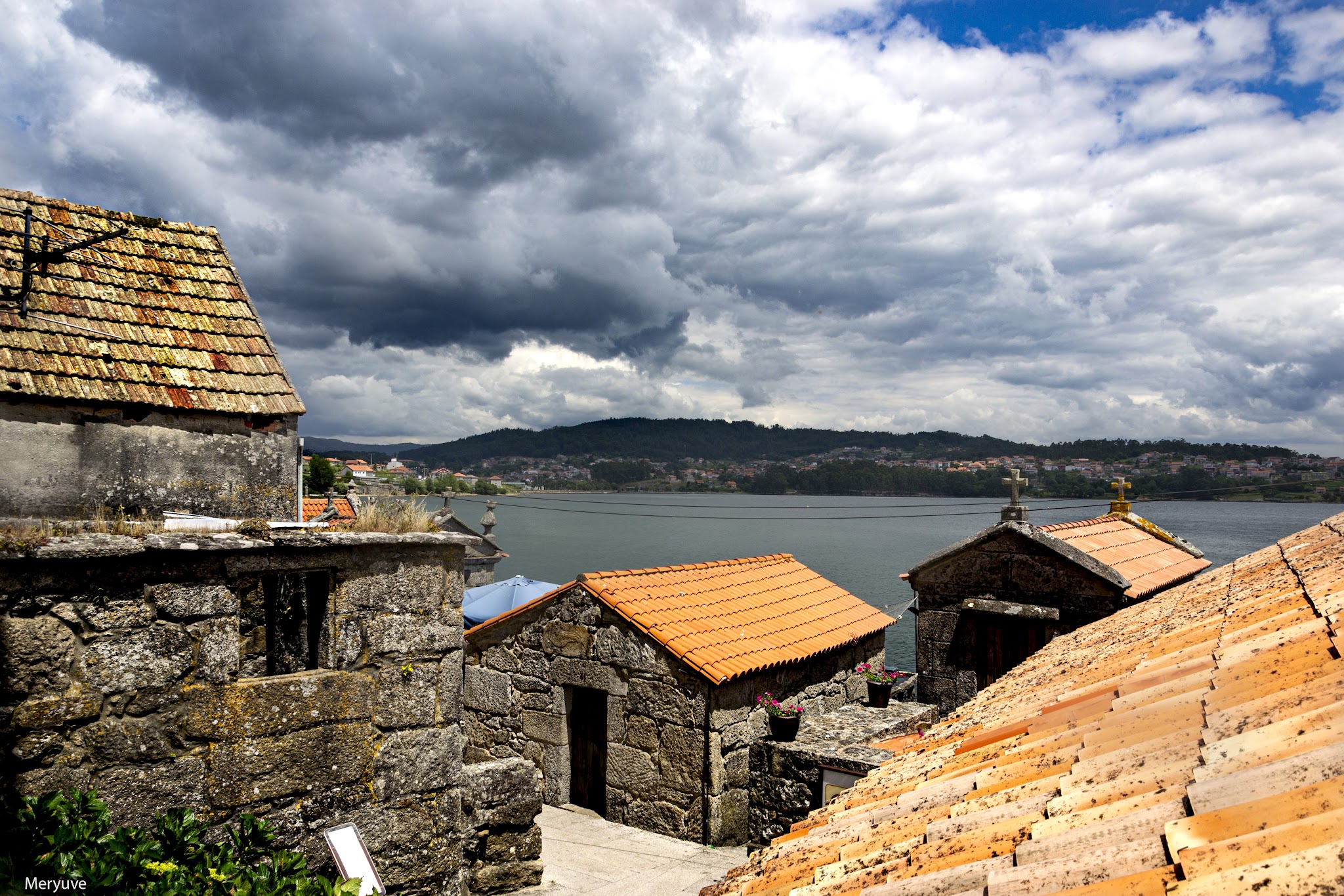 Steinhäuser mit orangefarbenen Ziegeldächern überblicken eine Bucht unter dramatischem Wolkenhimmel.