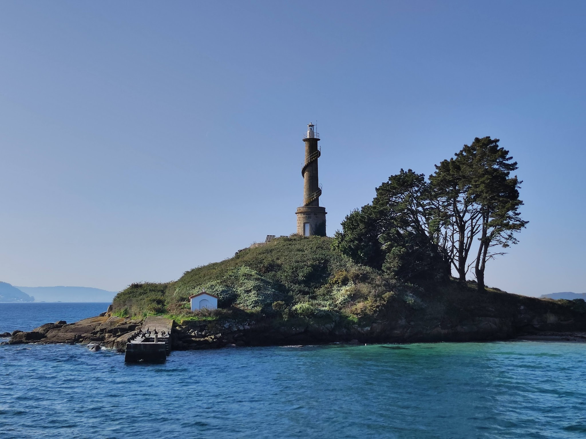 Steinleuchtturm mit äußerer Wendeltreppe auf grüner Insel, daneben Bäume und Steg im klaren blauem Meer.