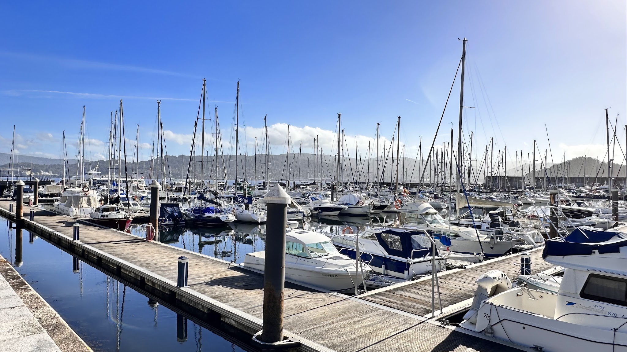 Sonnige Marina mit dicht aneinandergereihten Segelbooten und Yachten an Holzstegen, ruhiges Wasser, Berge im Hintergrund