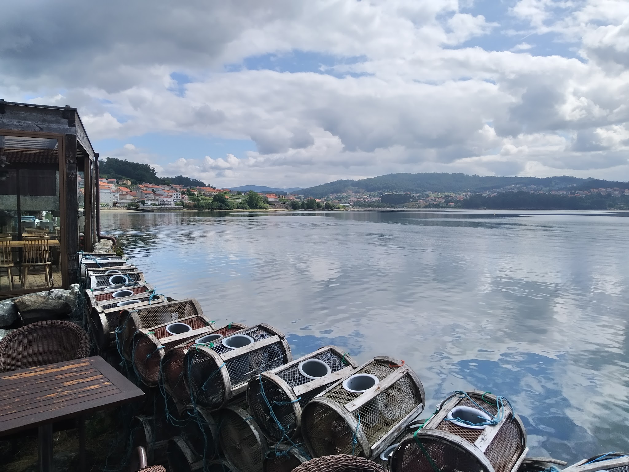 Stacked lobster traps beside a lakeside restaurant; calm water reflects clouds, distant hillside village.