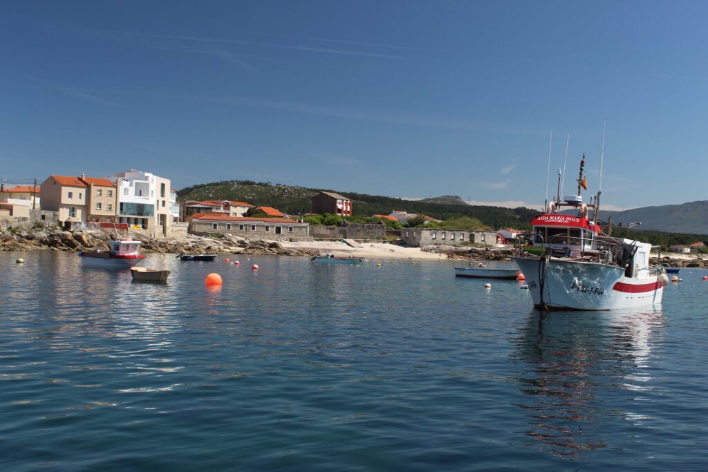 Kleines Küstendorf mit orange gedeckten Häusern; Fischkutter liegt auf ruhigem, blauem Wasser zwischen Bojen.