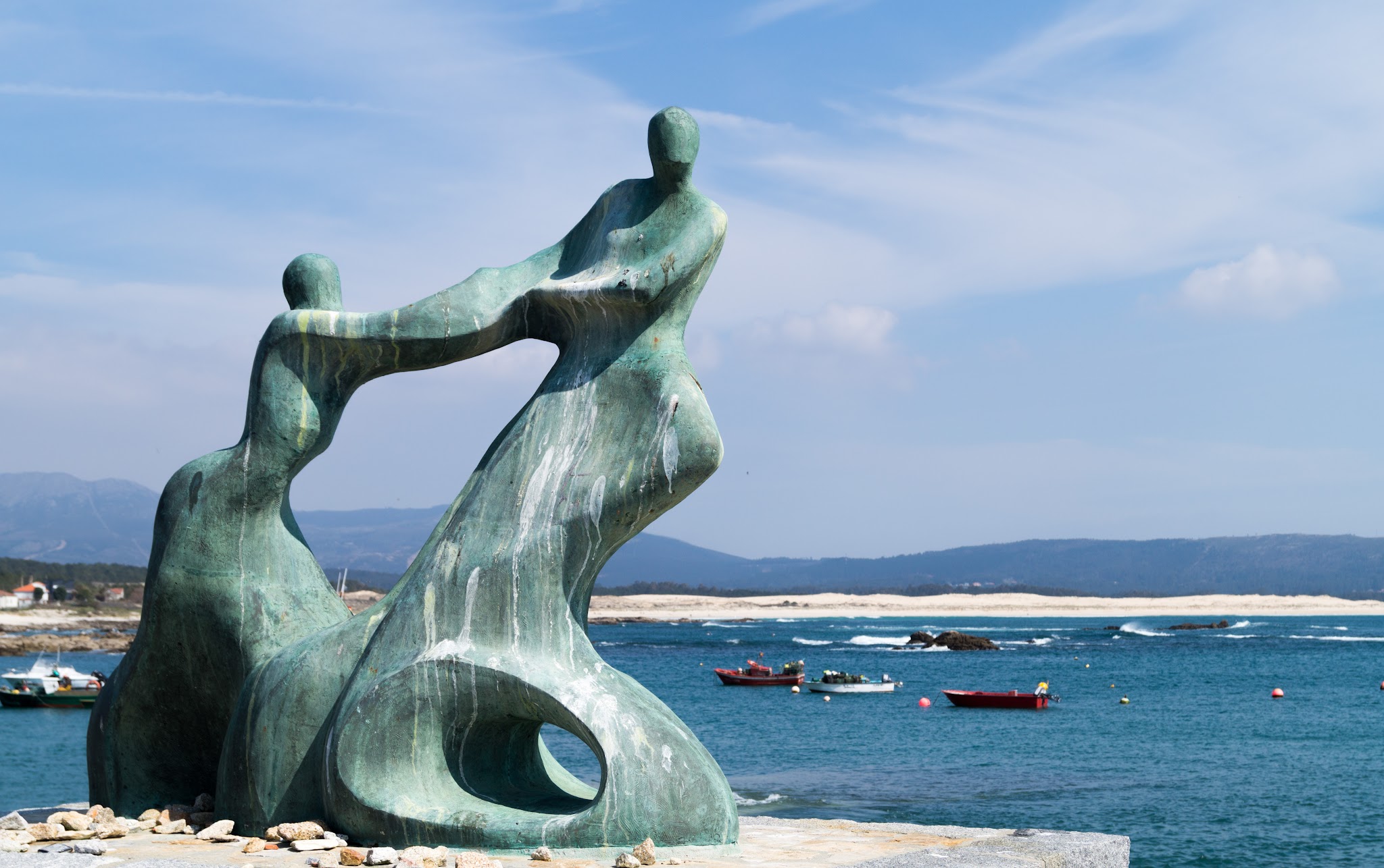 Abstrakte Bronze-Skulptur zweier sich haltender Figuren am Meeresufer; Boote und Strand im Hintergrund.