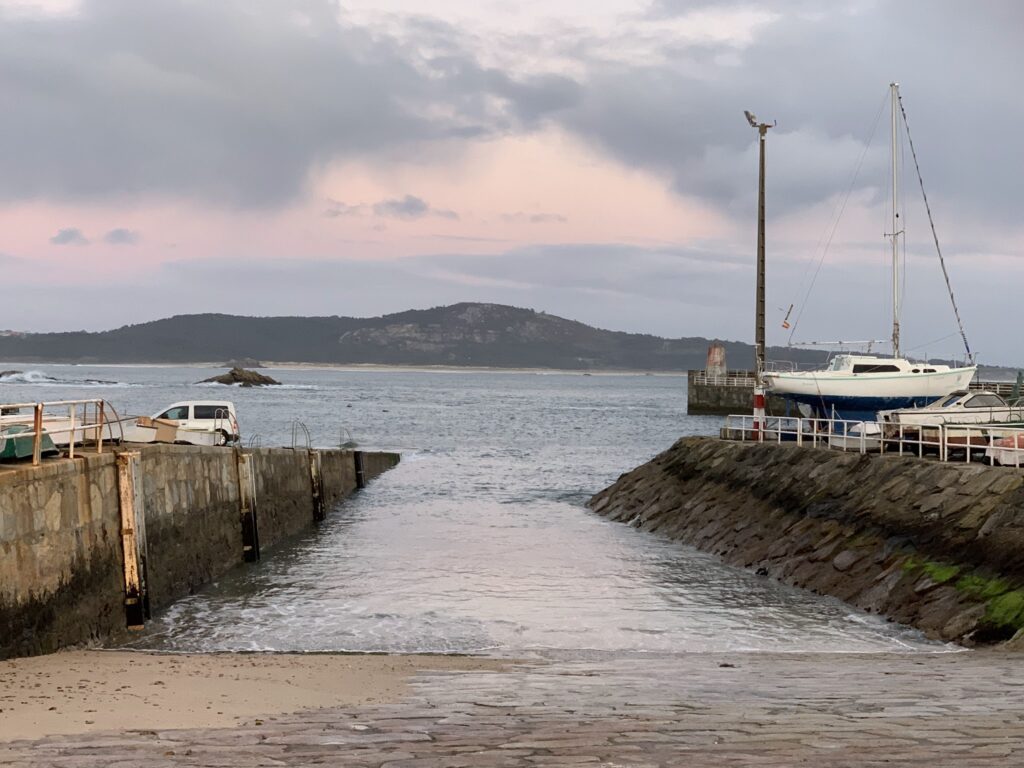 Slipanlage zwischen Steinmauern, rechts hochgebocktes Segelboot, ruhiges Meer vor Hügeln im rosa Abendlicht.