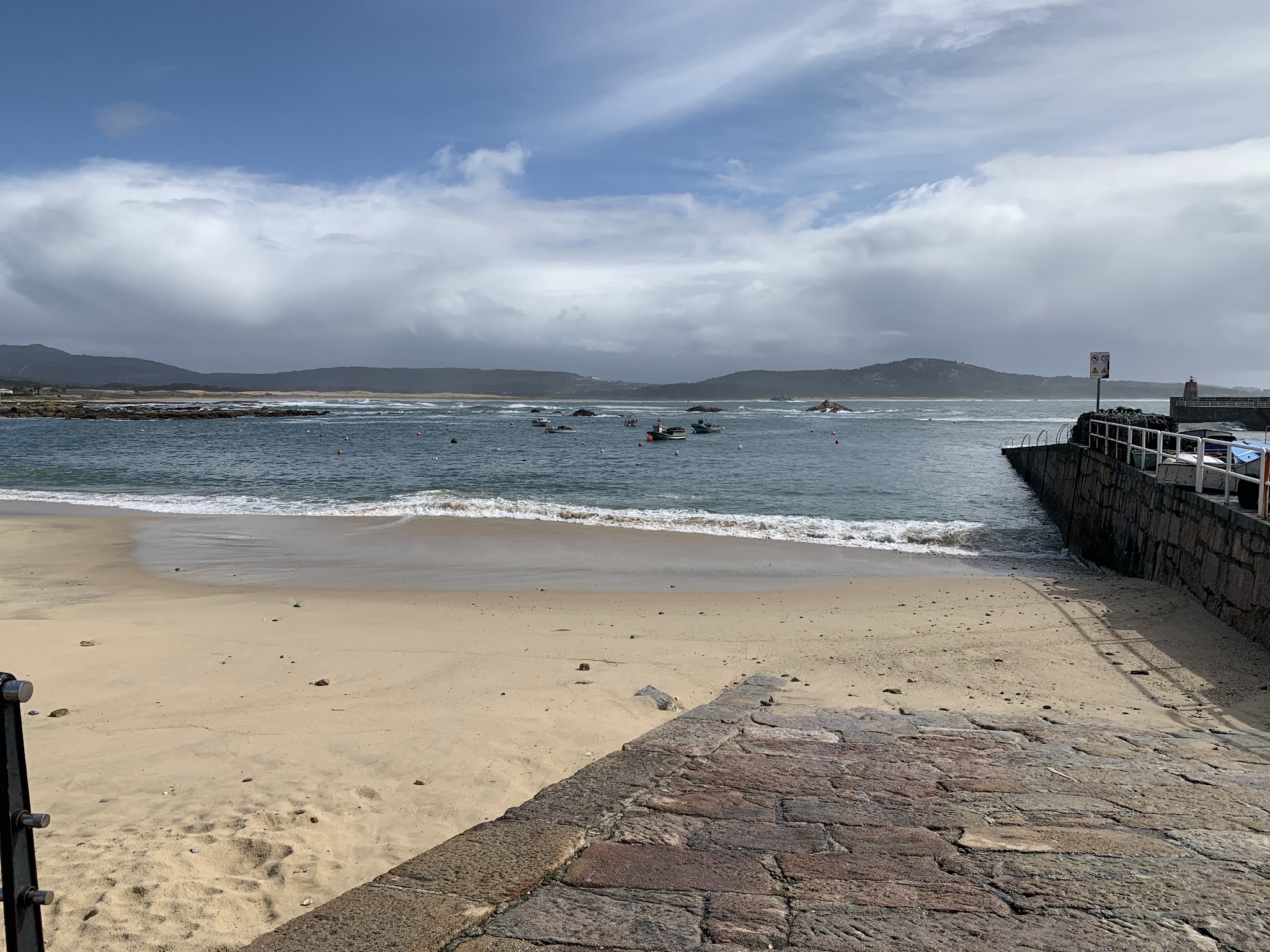 Breiter Sandstrand, wenige Boote in ruhiger Bucht, Steinpier rechts, bewölkter Himmel über fernen Hügeln