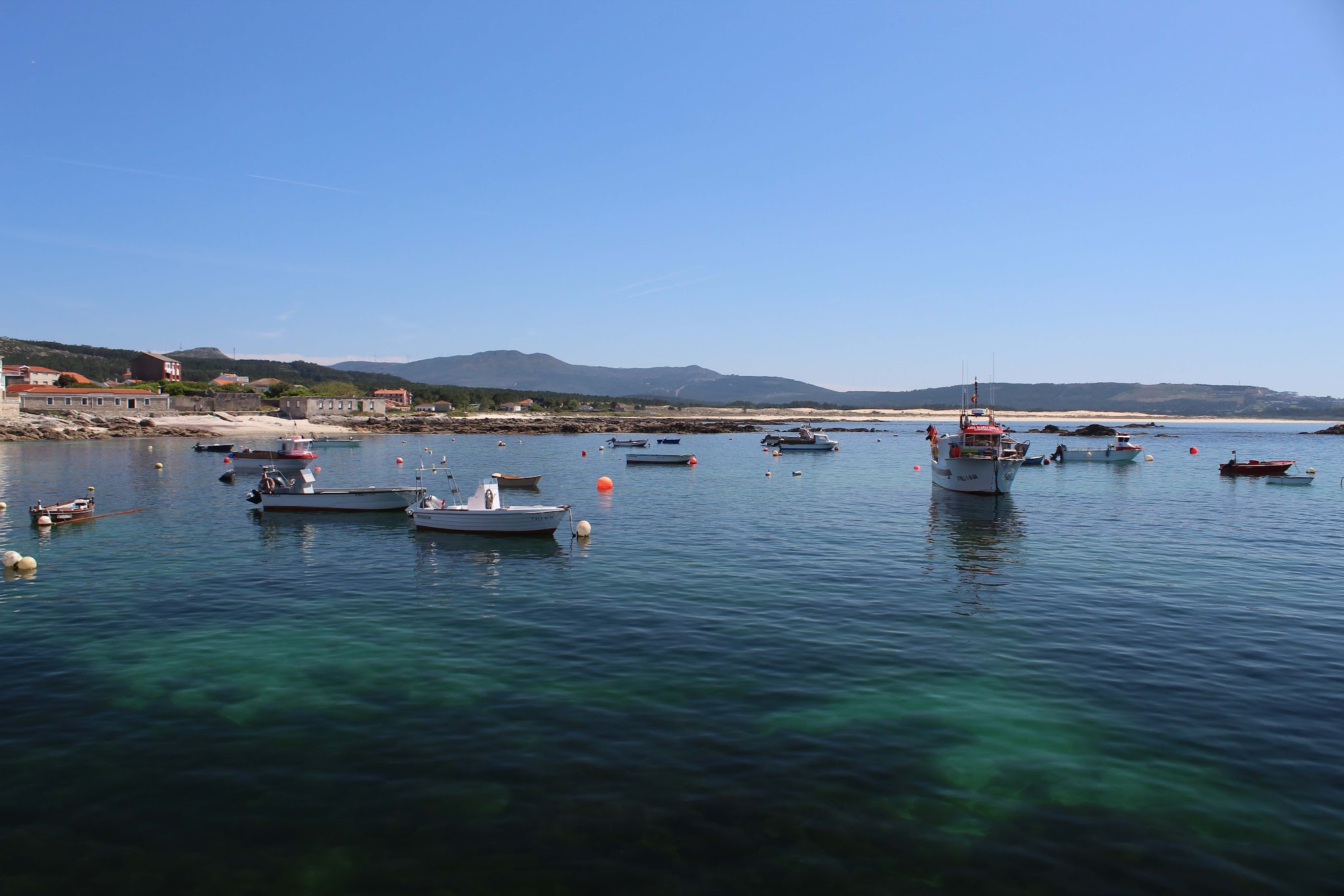 Mehrere Fischerboote liegen auf ruhigem, türkisblauem Meer vor einem kleinen Küstendorf mit Hügelkulisse.