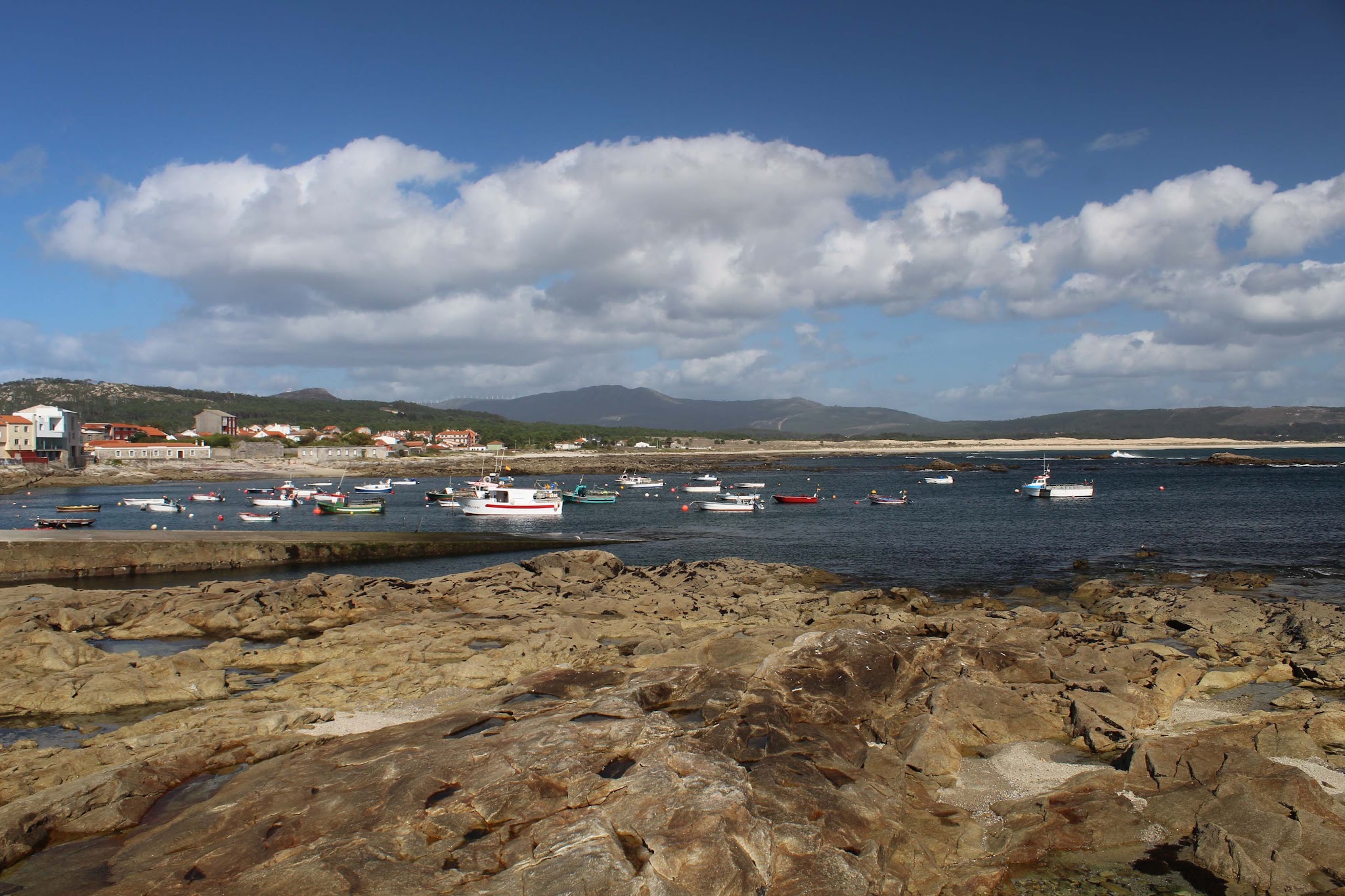 Kleine Fischerboote liegen vor einem Küstendorf; im Vordergrund Felsen, im Hintergrund Berge und Wolkenhimmel.