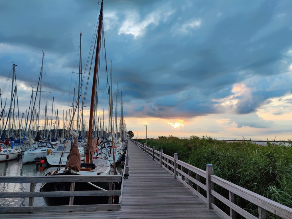 Segelboote liegen am Anleger, ein Holzsteg führt zwischen Hafen und Schilf ins Abendlicht unter dramatischem Wolkenhimmel.
