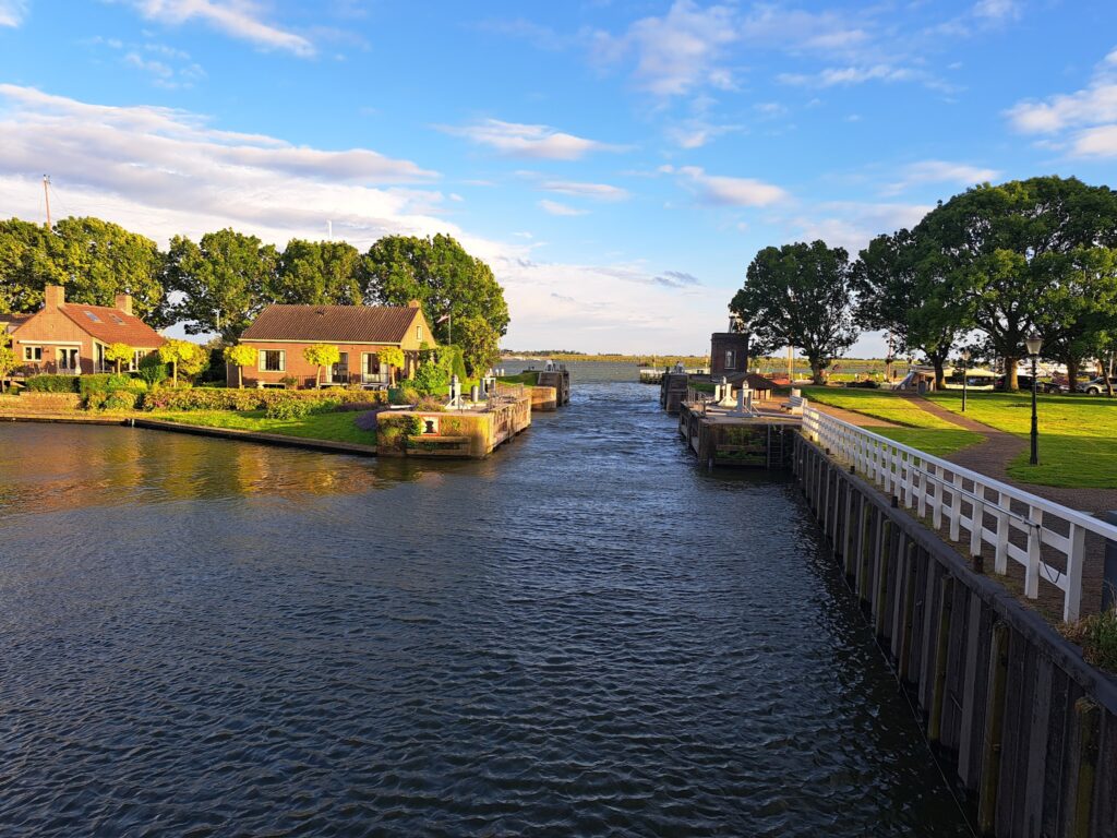 Sonniger Kanal mit Schleuse, Backsteinhäusern, grünen Bäumen und Blick aufs offene Wasser unter blauem Himmel.
