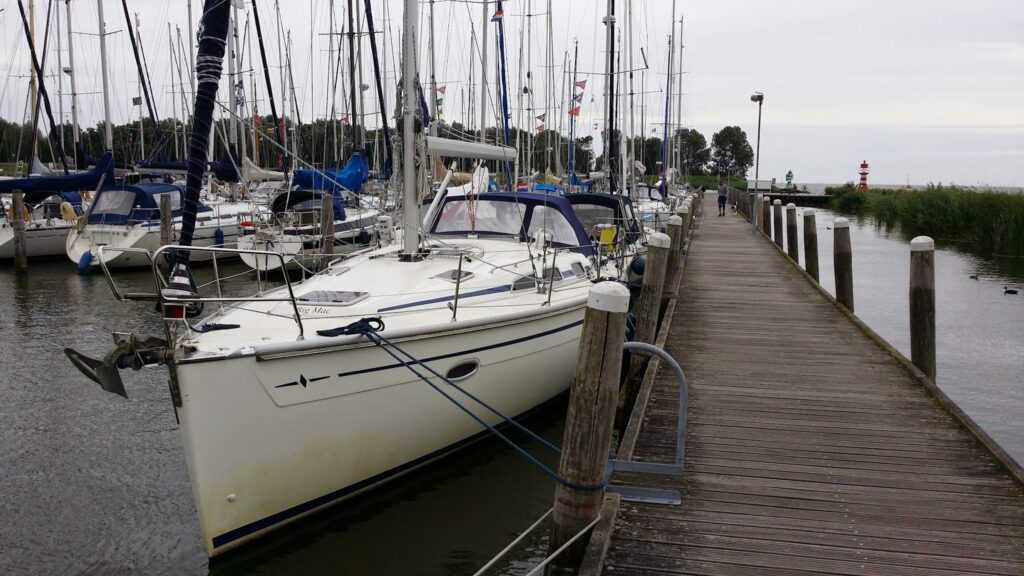 Segelyachten liegen längs eines hölzernen Stegs; Person geht in Ferne, kleiner roter Leuchtturm am Horizont.