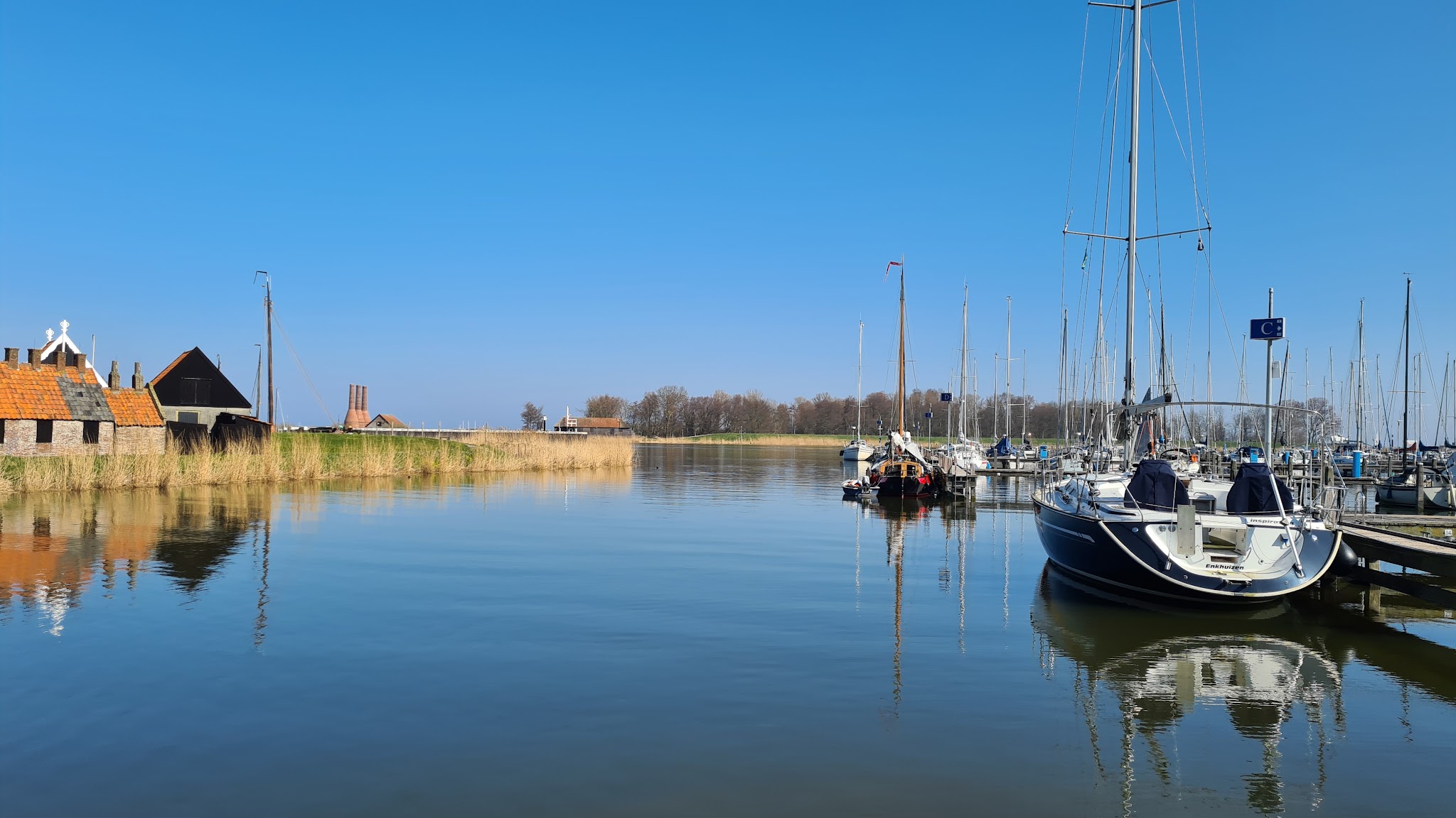 Ruhiger Hafen: Segelboote spiegeln sich im stillen Wasser, Schilf und alte Backsteinhäuser unter wolkenlosem Himmel